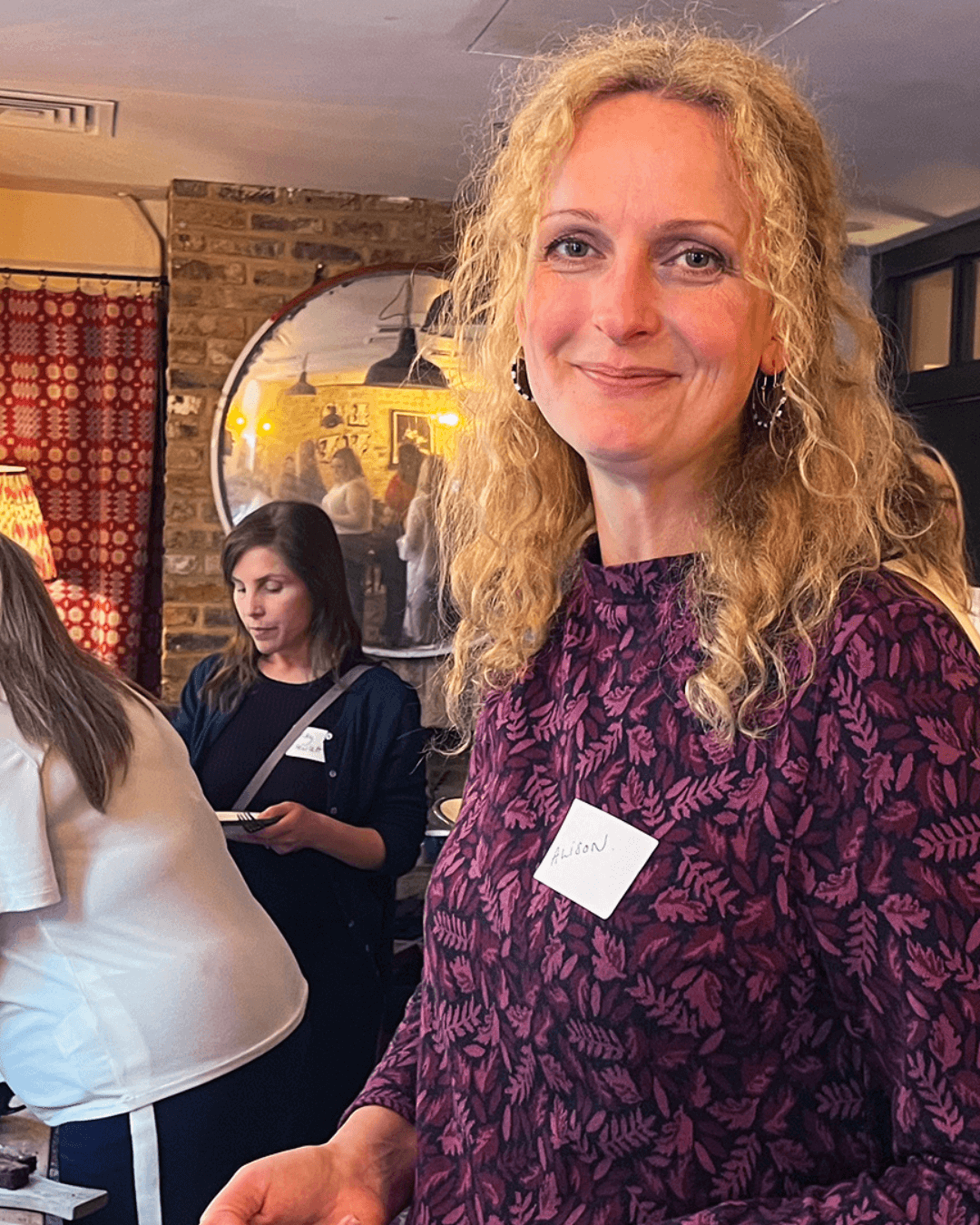 Shows a pretty blond woman standing in the middle of a vibrant event, she is smiling and looking happy. Around her other women are chatting and eating delicious snacks