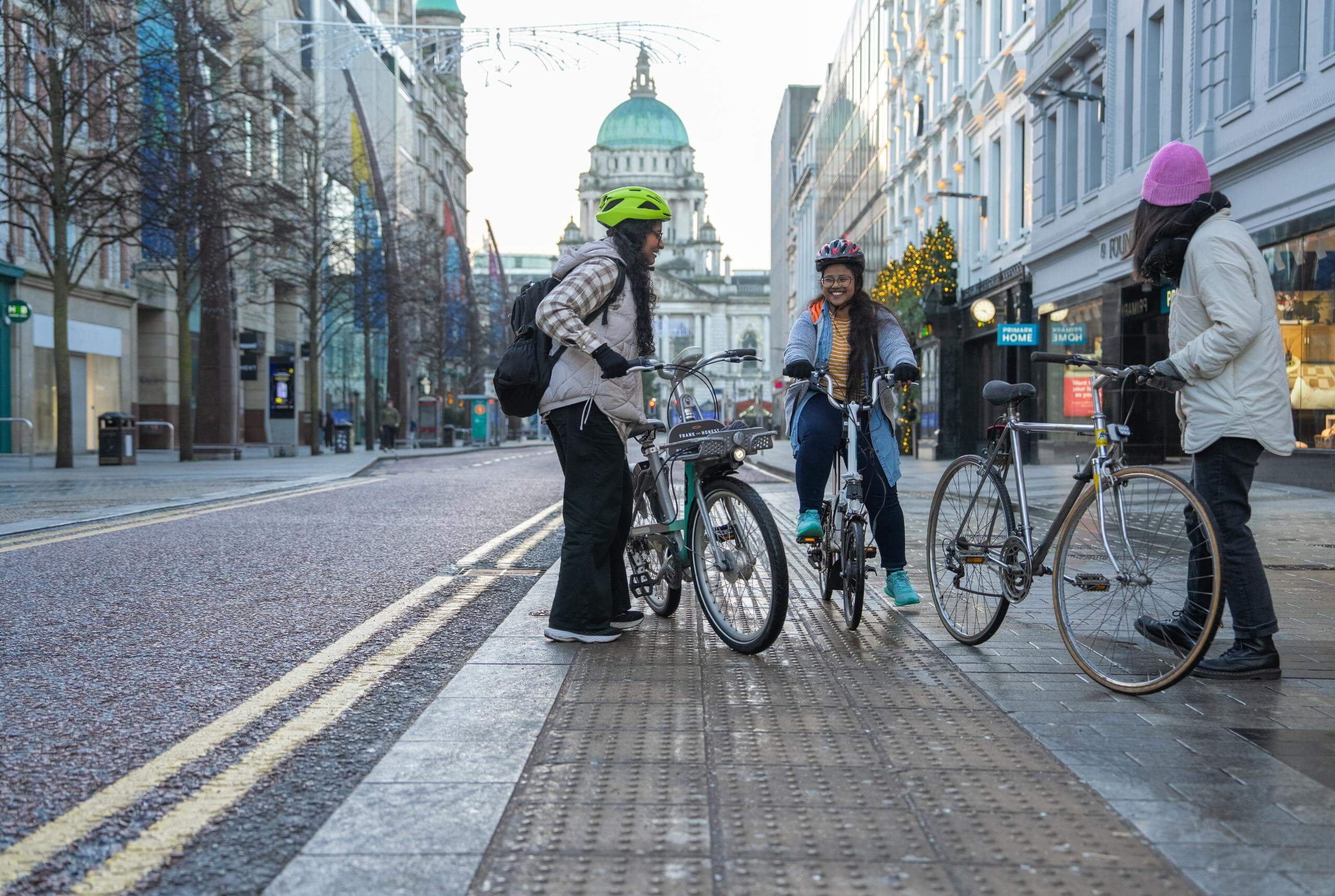 Three young women gather outside famous buildings in Belfast, they're astride bikes and wearing winter clothes. They are having fun.