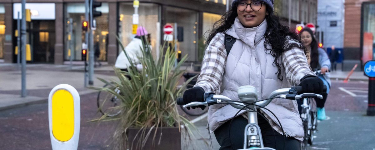 A woman on a bike cycling along in the middle of a city, she is wearing winter clothes and has brown skin, glasses and long brown hair, she's smiling.