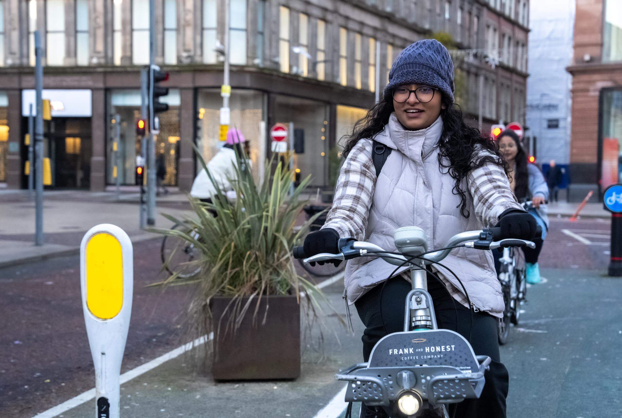 A woman on a bike cycling along in the middle of a city, she is wearing winter clothes and has brown skin, glasses and long brown hair, she's smiling.