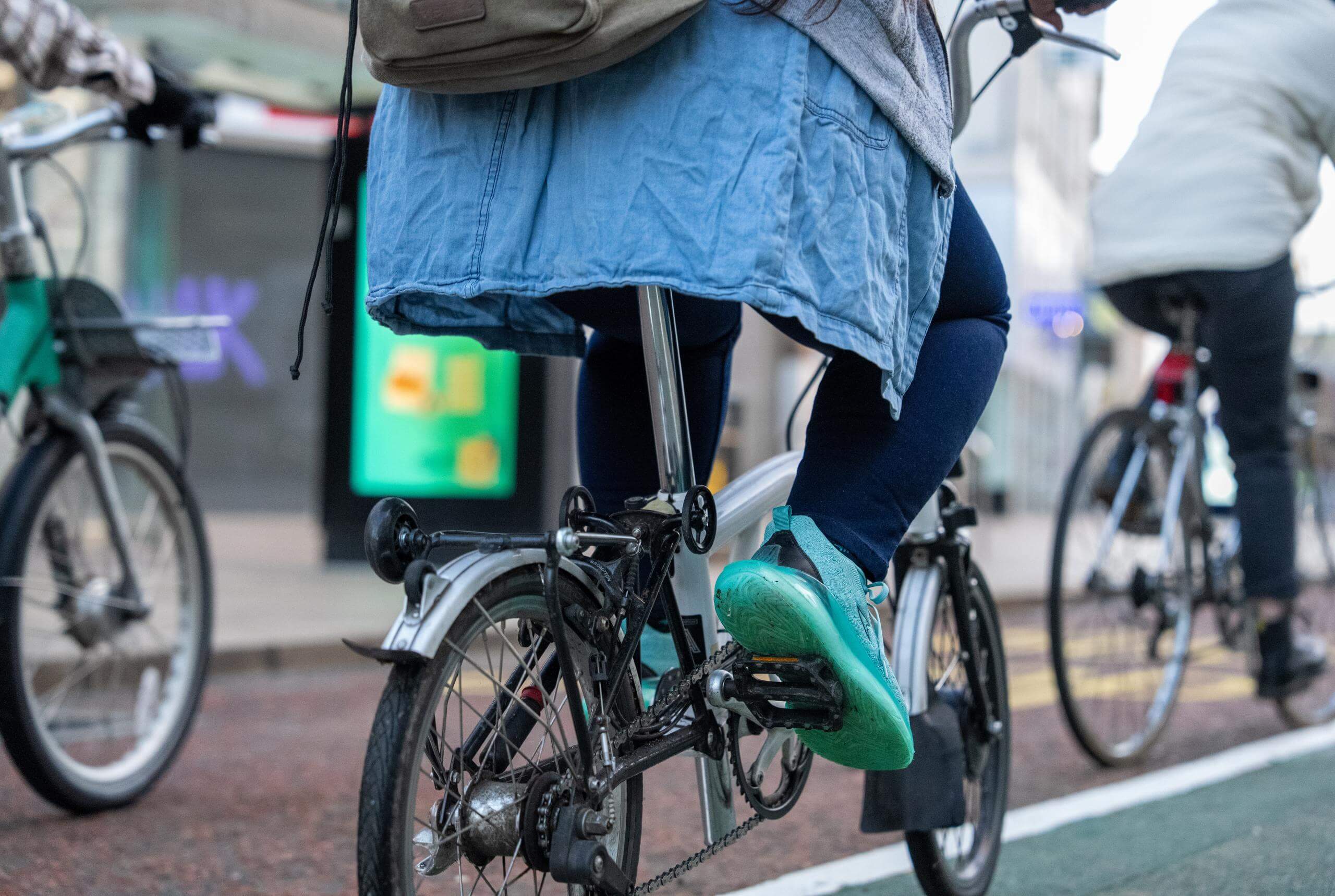 A close up of a bike, showing the back wheel with a woman riding, she is wearing bright green trainers and a blue coat