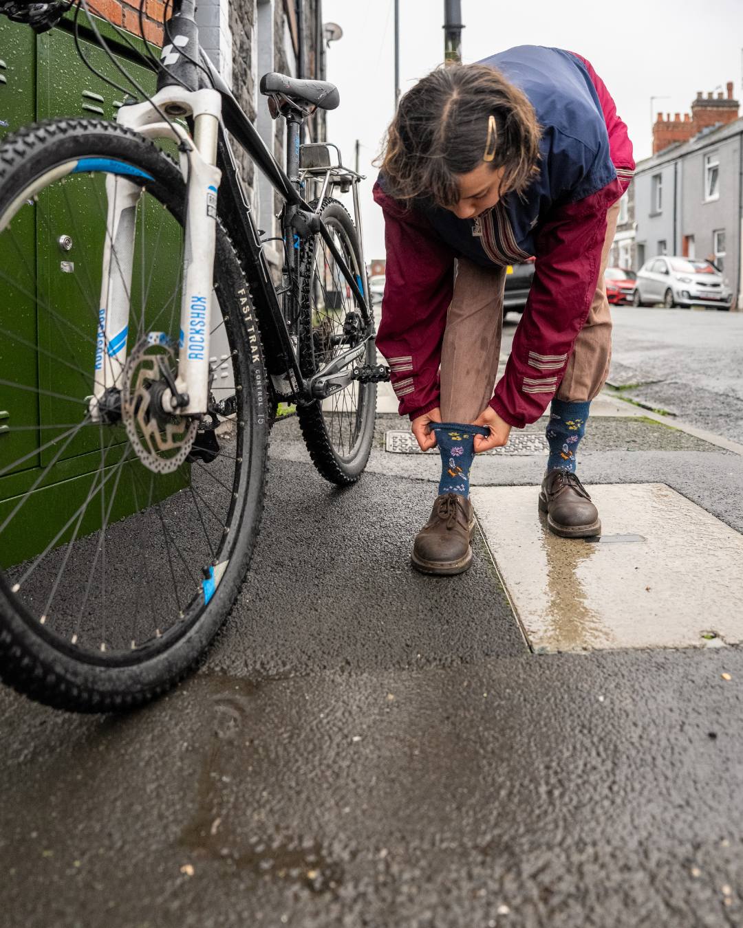 A young woman stands next to her bike in the street, she's bending down to pull her socks up over her trousers