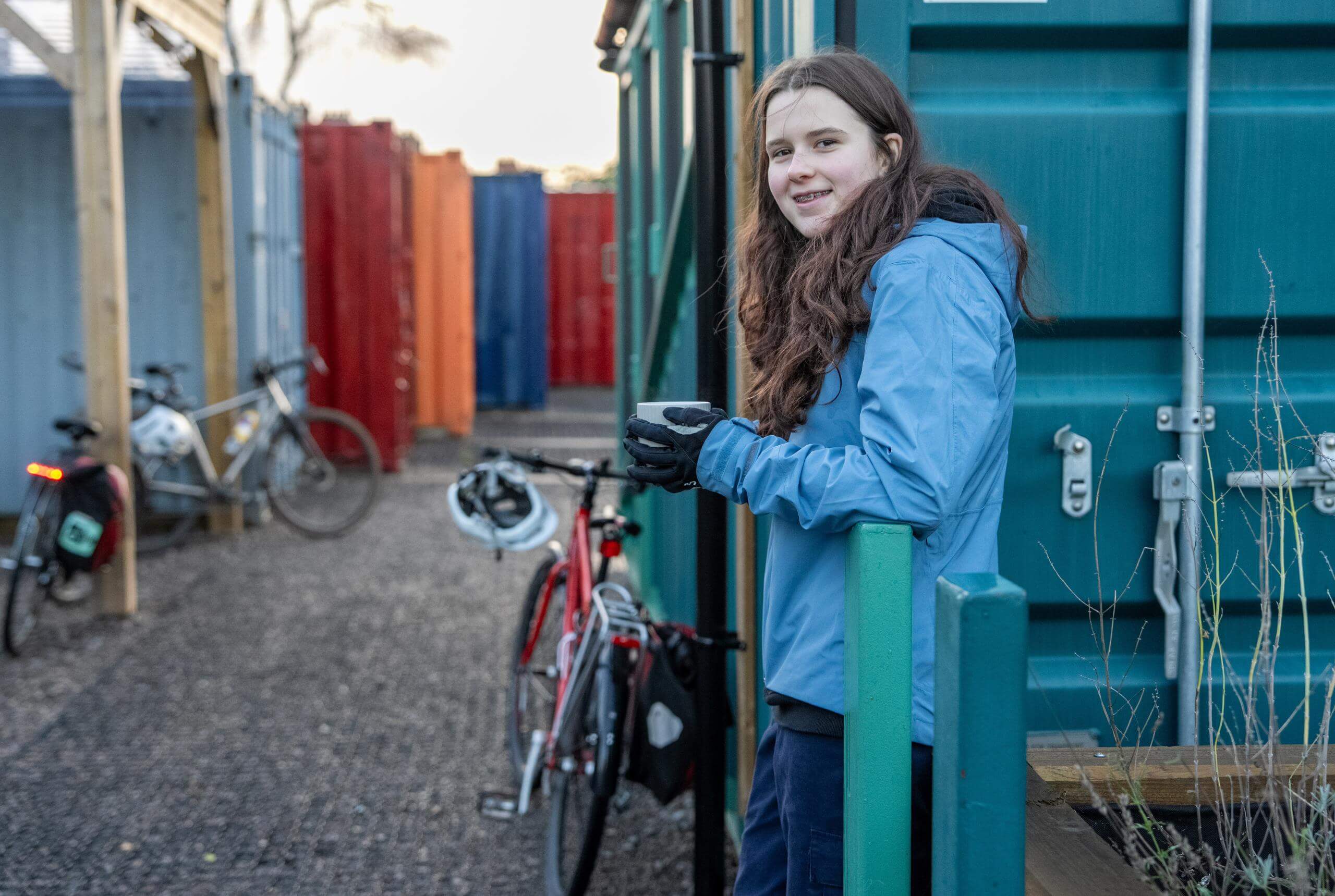 A young woman drinking a cup of tea outside, she's leaning on a brightly coloured storage container, her bike and helmet are leaning alongside her. It's winter, she's wearing a blue waterproof coat and has long brown hair. She's smiling
