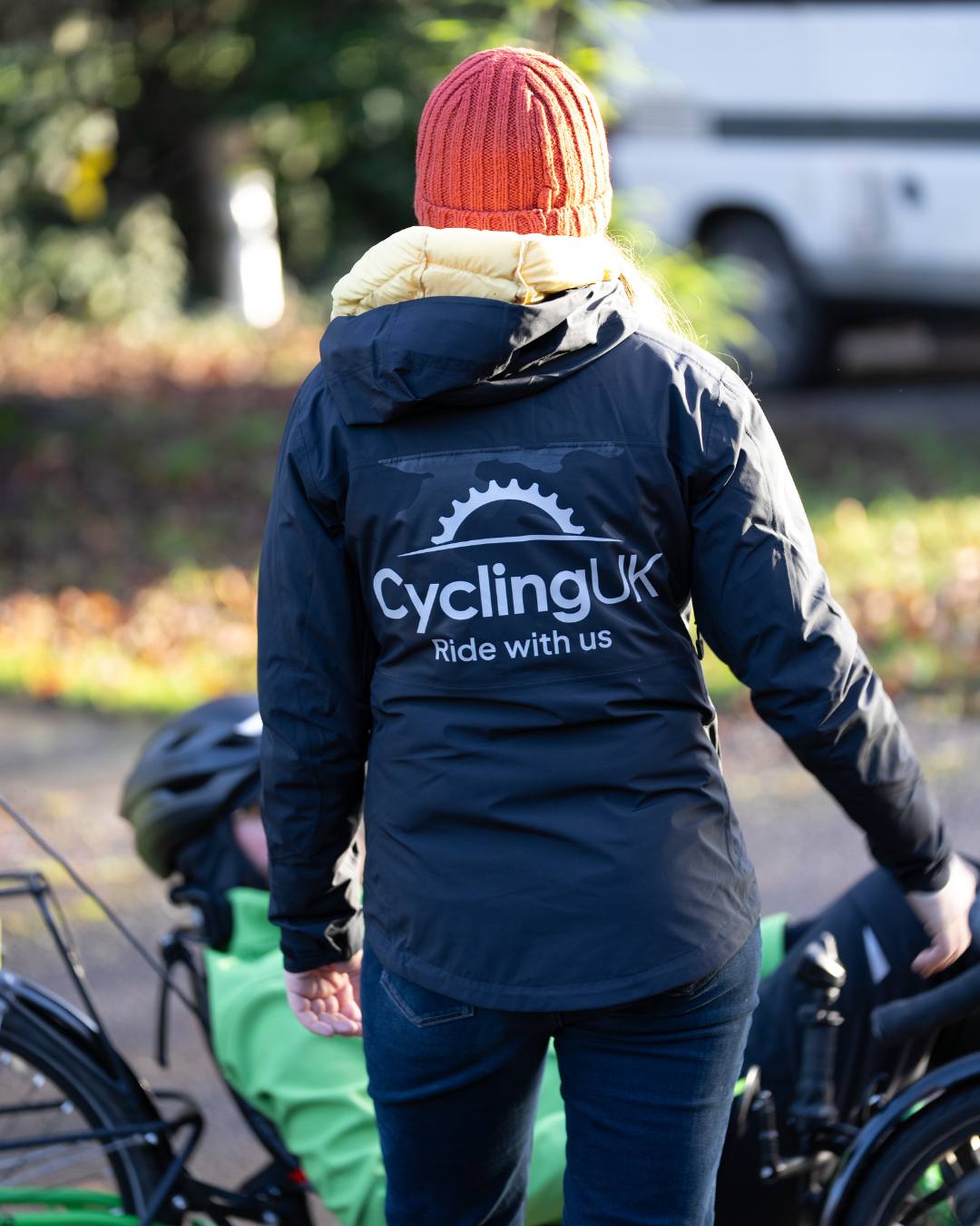 A woman turned with her back to the camera, its a sunny but cold day. She's wearing an orange hat and a blue coat with the Cycling UK logo on the back