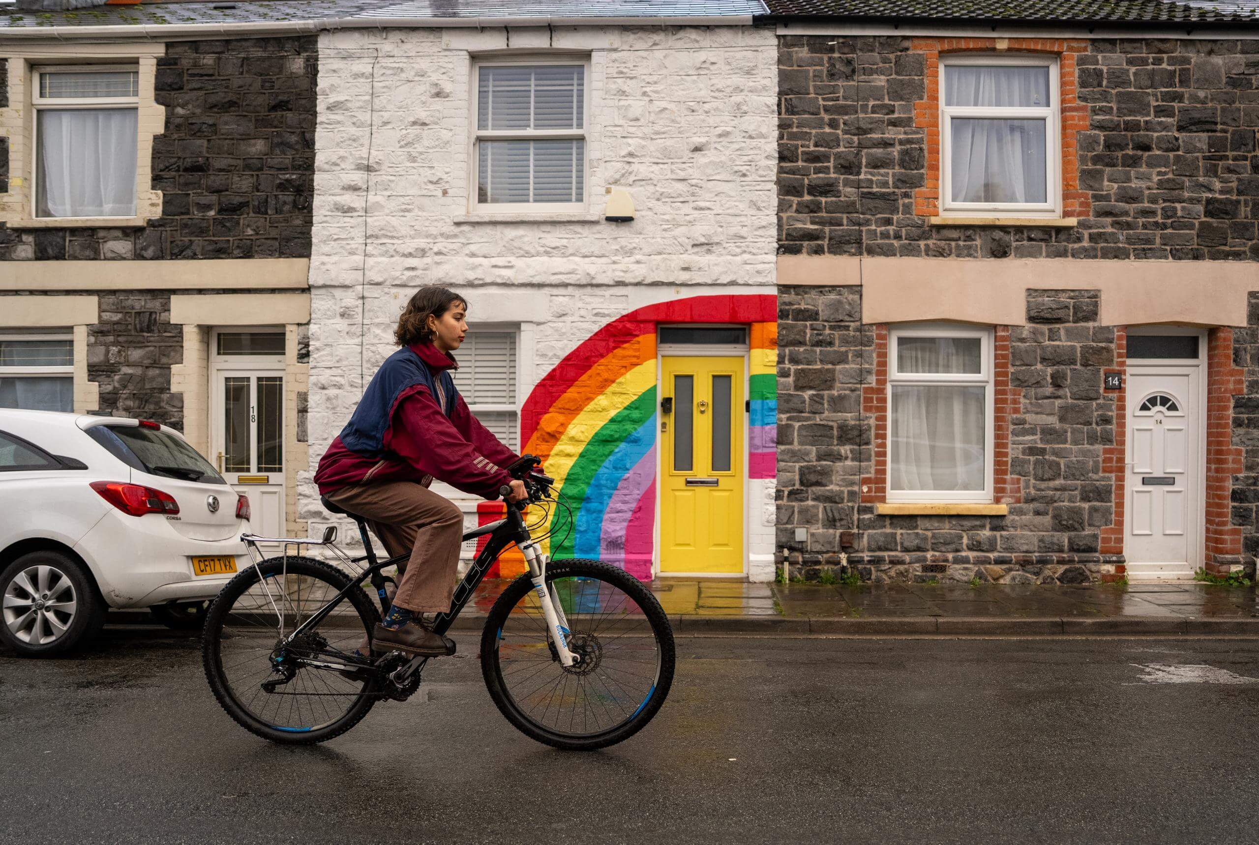 A young woman seen from the side as she cycles past a row of terraced houses, one is painted white with a rainbow and a yellow door.
