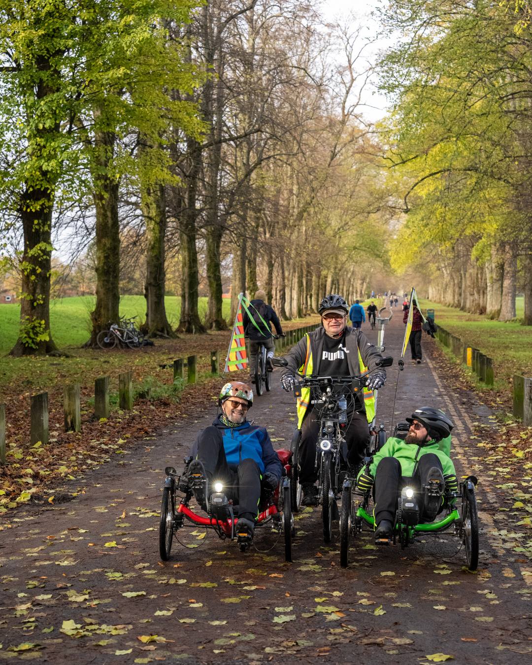Three men on bikes - one upright, two recumbent pose on a path through a park on a sunny autumn day. Autumn leaves are underfoot and on the trees overhead