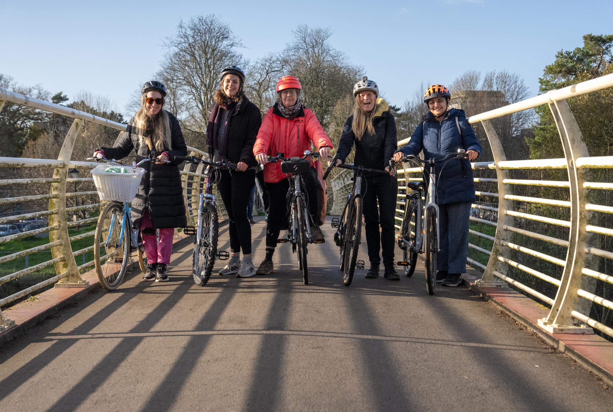 Five middle aged women stand astride their bikes on a modern bridge in a park. The sun is shining, it's winter and they're wearing waterproof coats and helmets, they look happy
