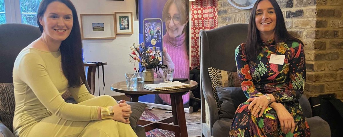 Laura Mitchell of SSAFA and Abi Mellor of MOREVER are sitting in chairs smiling at the camera, at the MOREVER Meetup. They both have long brown hair. Laura is wearing a green dress and Abi is wearing a floral dress. Behind them is the Give A Little banner.