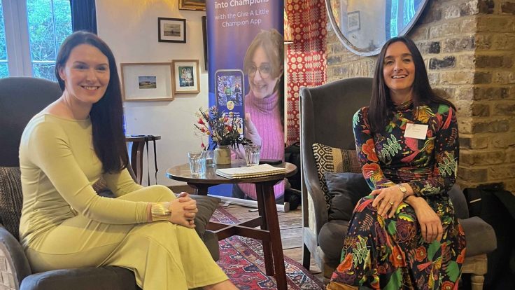 Laura Mitchell of SSAFA and Abi Mellor of MOREVER are sitting in chairs smiling at the camera, at the MOREVER Meetup. They both have long brown hair. Laura is wearing a green dress and Abi is wearing a floral dress. Behind them is the Give A Little banner.