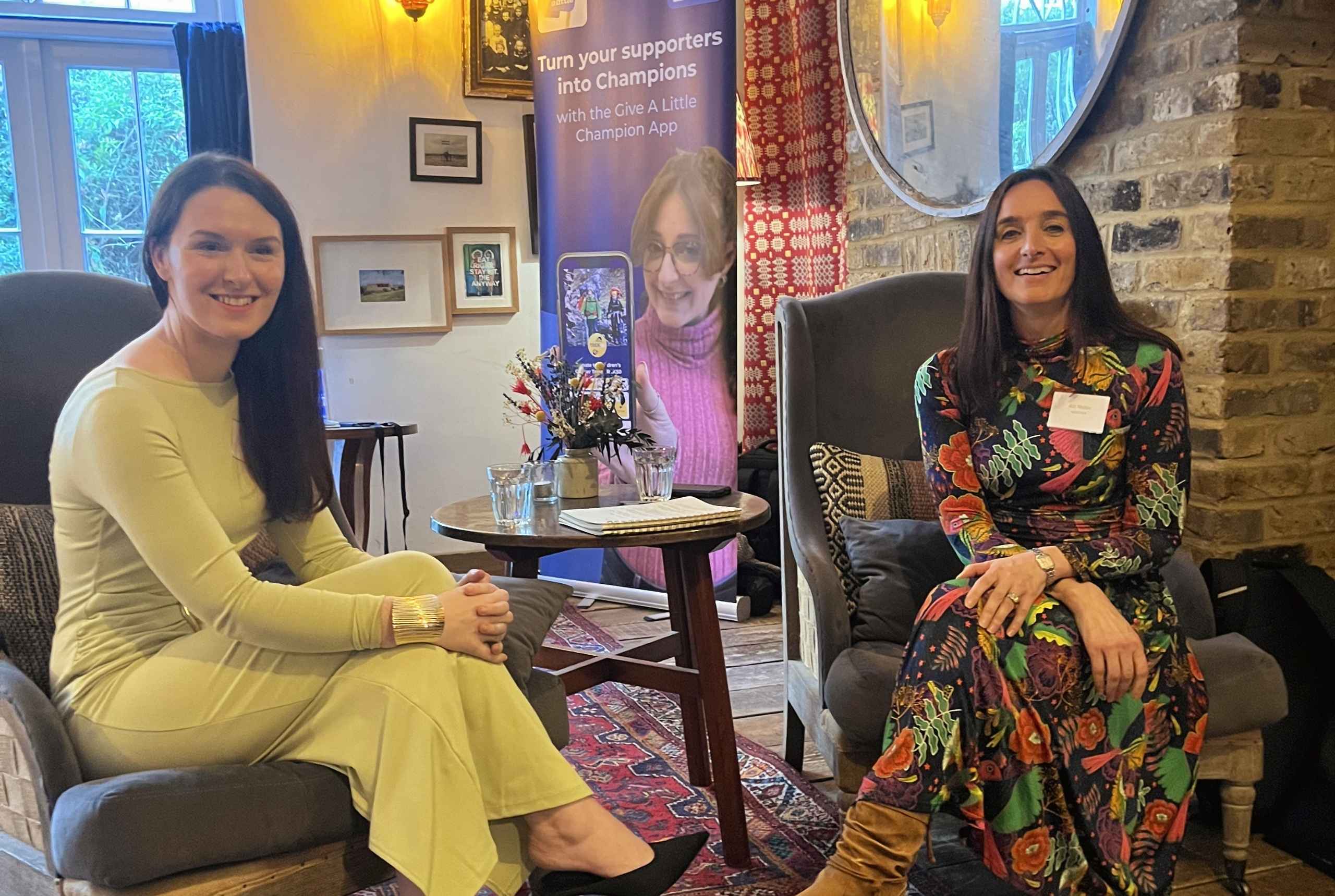 Laura Mitchell of SSAFA and Abi Mellor of MOREVER are sitting in chairs smiling at the camera, at the MOREVER Meetup. They both have long brown hair. Laura is wearing a green dress and Abi is wearing a floral dress. Behind them is the Give A Little banner.